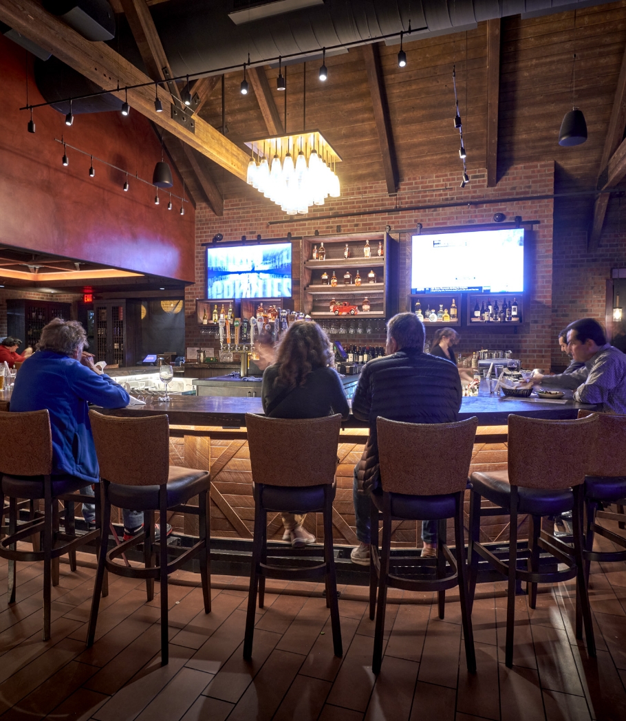 Restaurant guests seated at a bar, enjoying their time in a rustic setting with a backlit liquor shelf and televisions displaying sports.